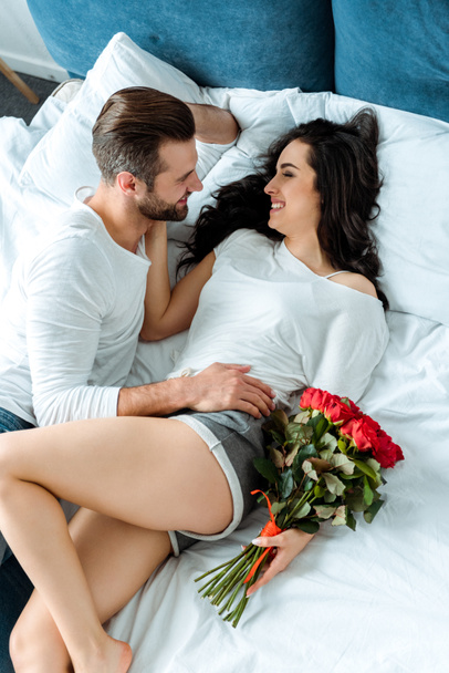 εναέρια θέα του ευτυχισμένο ζευγάρι ξαπλωμένο στο κρεβάτι με μπουκέτο κόκκινα τριαντάφυλλα - Φωτογραφία, εικόνα