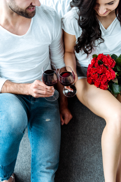 overhead view happy couple clinking with glasses of red wine and woman holding red roses in bedroom - Фото, изображение