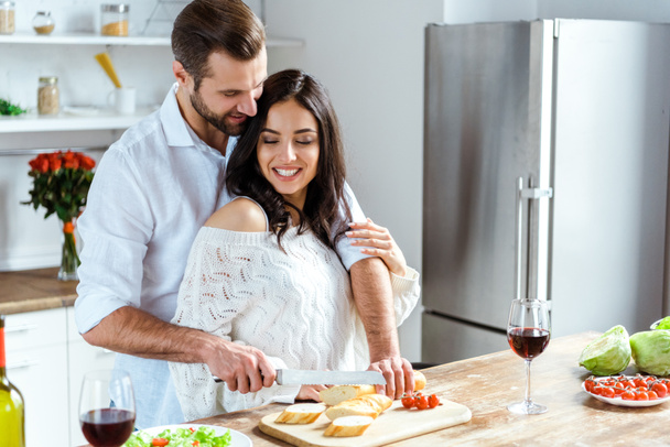 happy couple standing together at kitchen while man cutting bread on chopping board - Fotoğraf, Görsel