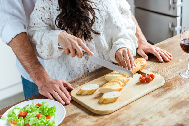 обрезанный вид пары, стоящей вместе на кухне, в то время как женщина режет хлеб на доске возле свежего салата
 - Фото, изображение
