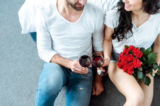 vista aérea de pareja sonriente tintineando con copas de vino tinto y mujer sosteniendo ramo de rosas rojas
 - Foto, Imagen