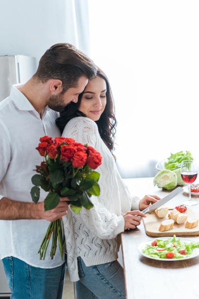 man holding bouquet of roses while embracing woman during cooking salad and cutting bread - Fotografie, Obrázek