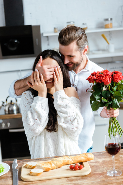 hombre sosteniendo ramo de rosas y cerrando ojos de mujer para hacer sorpresa en la cocina
 - Foto, imagen