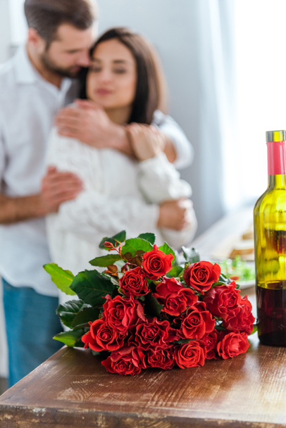 selective focus of man embracing woman near bouquet of red roses and bottle of wine - Foto, imagen