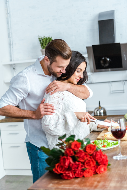 赤いバラの花束の近くのキッチンでロマンチックなディナーを調理中に女性を抱きしめる男 - 写真・画像