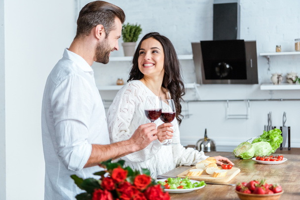 happy man clinking glasses of red wine with smiling woman at kitchen - Foto, afbeelding