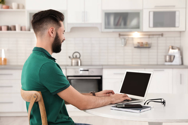 Handsome young man working with laptop at table in kitchen - Photo, image