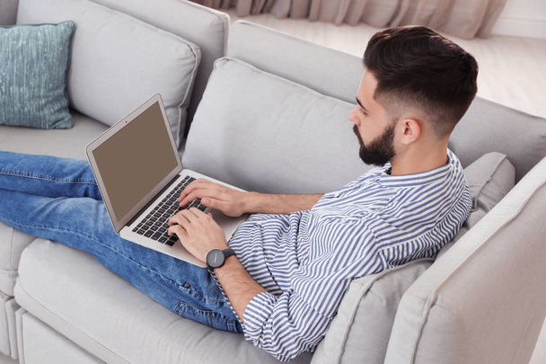 Joven guapo trabajando con el ordenador portátil en el sofá en casa
 - Foto, Imagen