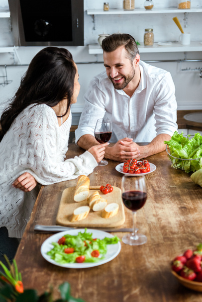 fiatal pár iszik vörösbort fa asztalnál cseresznye paradicsom, kenyér és saláta - Fotó, kép