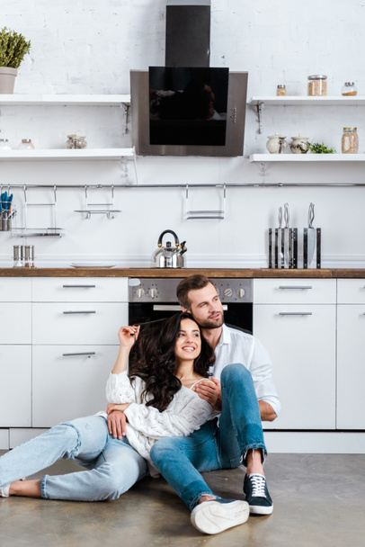 happy couple sitting on floor and hugging at kitchen and looking away - Foto, Imagem