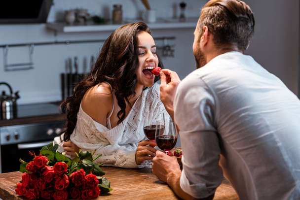 coppia con bicchieri di vino rosso a tavola in legno con bouquet di rose, donna che alimenta l'uomo con fragola
 - Foto, immagini