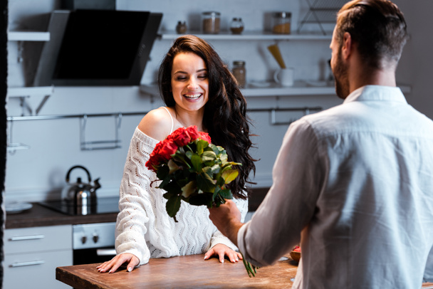 man gifting bouquet of roses to smiling woman at kitchen - Fotó, kép