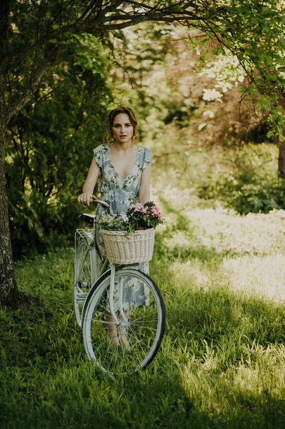 portrait of a beautiful girl in the forest, holding a bike with a basket of flowers, behind the rays of the sun, a blue flowered dress, summer walk - Photo, image