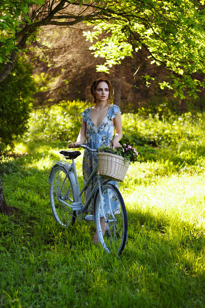 portrait of a beautiful girl in the forest, holding a bike with a basket of flowers, behind the rays of the sun, a blue flowered dress, summer walk - Foto, afbeelding