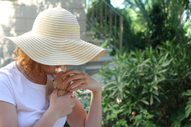 Teenager-Mädchen mit Strohhut hält an einem sonnigen Tag einen kleinen Teddybär im Garten - Foto, Bild