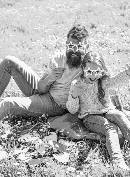 Father and daughter sits on grass at grassplot, green background. Child and dad posing with star shaped eyeglases photo booth attribute at meadow. Family spend leisure outdoors. Superstar concept - 写真・画像