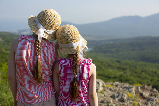 Back view of two little cute girls in lovely summer straw caps with lace ribbons are standing together and looking at the beautiful landscape in mountains near Ghost valley and the Black sea, Crimea. - Foto, Bild