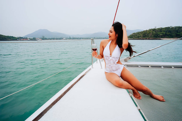 Young happy Asian woman enjoy and hold a wineglass sitting on deck of sailing yacht boat in bikini - Foto, afbeelding