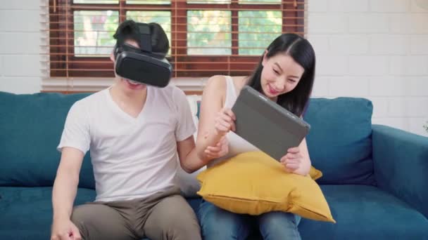 Coppia asiatica che utilizza tablet e simulatore di realtà virtuale che gioca in salotto, coppia sentirsi felici utilizzando il tempo insieme sdraiati sul divano a casa. Stile di vita famiglia relax a casa concetto
. - Filmati, video