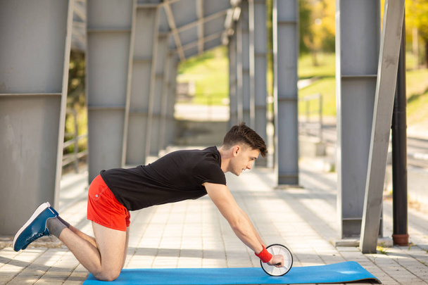 Young man training yoga outdoors. Sporty guy makes stretching exercise on a blue yoga mat, on the sports ground. - Foto, afbeelding