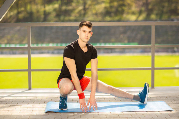 Молодой человек тренирует йогу на открытом воздухе. Спортивный парень делает упражнения на растяжку на синем коврике для йоги, на спортивной площадке
. - Фото, изображение
