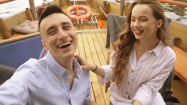 Young man and woman cheerfully doing selfie on ship in open air. Selfie video - Romantic couple taking selfie video by sunset over the river on small cruise ship Woman and man taking cell phone photos - Felvétel, videó