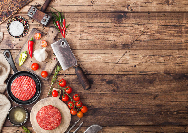 Fresh raw minced homemade farmers grill beef burgers on round chopping board and frying pan with spices and herbs and meat hatchet on wooden board. Oil, garlic and tomatoes. - Fotoğraf, Görsel