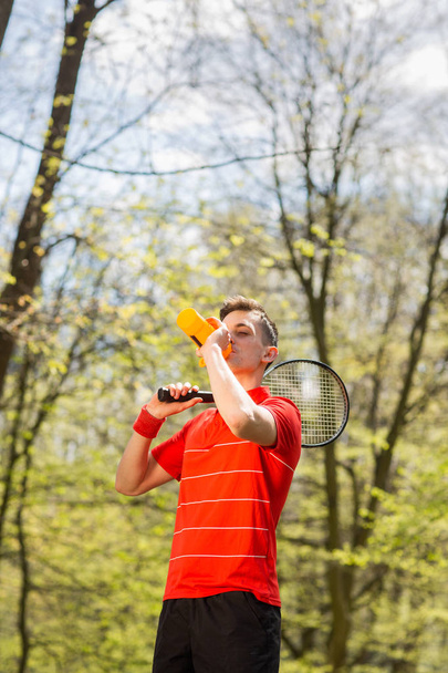Człowiek w czerwonym t-shirt stanowią z rakieta tenisowa i wody pitnej z termopara. Koncepcja sportu - Zdjęcie, obraz