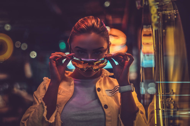 Attractive pensive styilish woman is posing for photographer in neon lights. There are darkness at background. - Fotó, kép