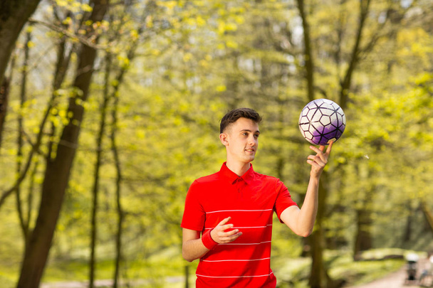 Kırmızı tişörtlü genç bir adam yeşil parkta futbol topuyla oynuyor. Spor konsepti - Fotoğraf, Görsel
