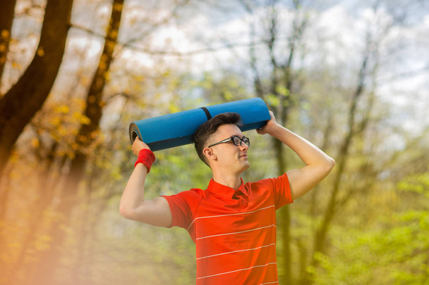 junger Sportler in rotem T-Shirt posiert in einem Frühlingspark mit einer blauen Yogamatte. er hält sich eine Yogamatte über den Kopf - Foto, Bild