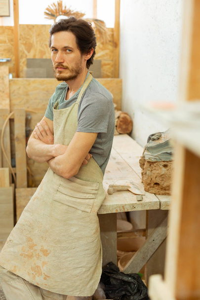Dark-haired bearded master in protective apron staying in workshop - Photo, Image