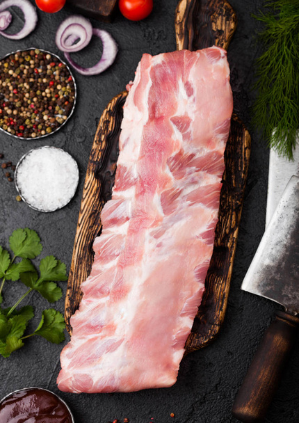 Fresh raw pork ribs on chopping board and vintage meat hatchets on black stone background. Fresh tomatoes and red onion with garlic salt and pepper.  - Foto, Imagen
