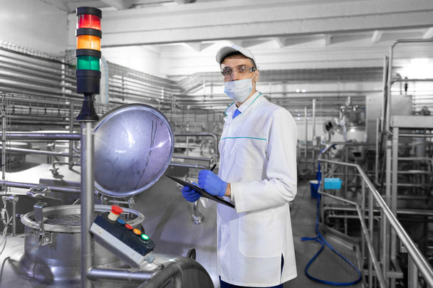 Инспектор в маске и скрабе стоит с папкой-планшетом в руках на молочном заводе
 - Фото, изображение