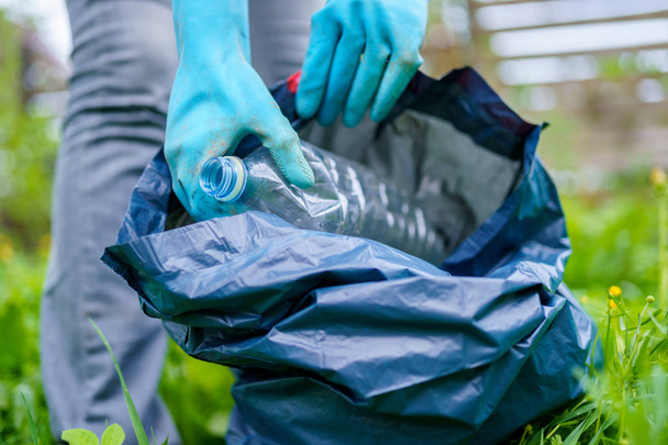 Photo of girl in rubber gloves picking up dirty plastic bottle in bag on green lawn - 写真・画像