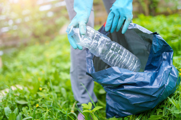 Photo of human in rubber gloves picking up dirty plastic bottle in bag on green lawn - Foto, imagen