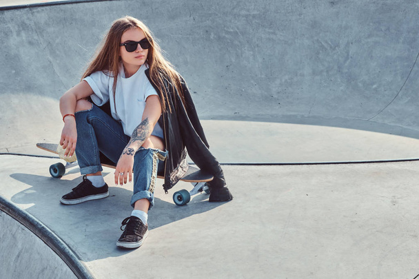Модна молода жінка з довгим волоссям і сонцезахисними окулярами сидить на скейтпарку
 - Фото, зображення