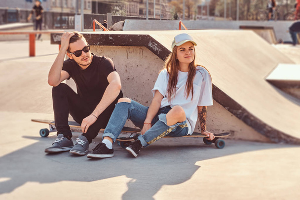 Joven pareja de moda están sentados en el parque de skate soleado con sus longboards
 - Foto, Imagen