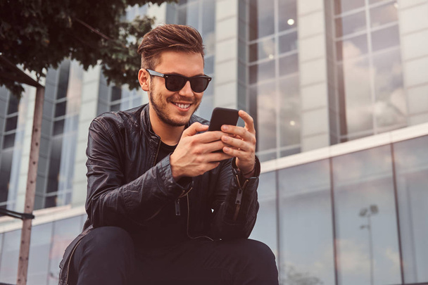 Усміхнений привабливий чоловік спілкується на мобільному телефоні, сидячи на вулиці біля будівлі
 - Фото, зображення