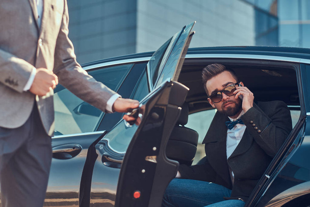 Busy businessman in sunglasses is talking by smartphone while his elegant assistant is opening door for him. - Foto, Bild