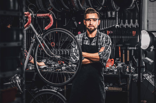 Ο όμορφος άντρας με γυαλιά στέκεται κοντά στο σταθερό ποδήλατο στο εργαστήριό του.. - Φωτογραφία, εικόνα