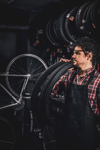 Pensive happy man in protective glasses is standing with tires at bicycle repairing studio - Foto, afbeelding