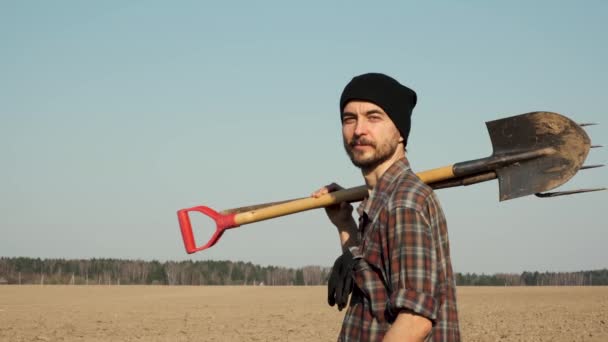 Portrait of smiling young caucasian bearded male farmer with shovel and pitchfork, stands on a freshly plowed field. Ready for spring farming work. - Кадры, видео