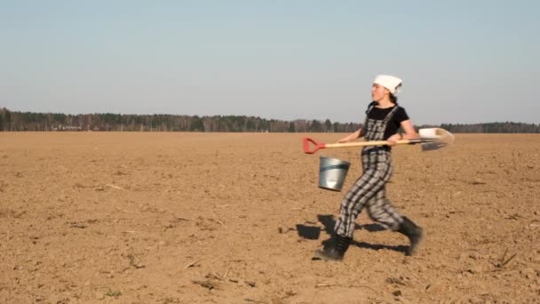 Happy life in countryside. Funny adult woman farmer with shovel and bucket jumps on freshly plowed field. Slow motion.  - Záběry, video