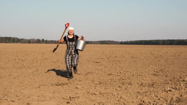 Funny cherrful woman farmer runs across ploughed field in jump throws to side shovel and bucket. Slow motion.  - Séquence, vidéo