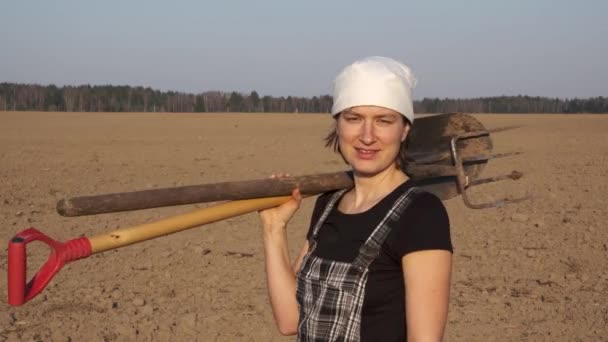 Portrait of woman farmer in worker coverall with tools on freshly plowed field on sunset. She turns, smiles, and walks away from the camera. - Кадри, відео