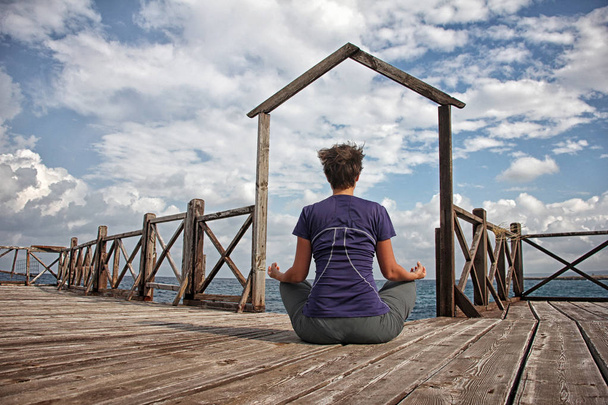 Gate to the sea. Athletic woman with short hair sitting in lotus pose on wooden pier and meditating against beautiful sky with clouds. View from behind.  - Fotó, kép
