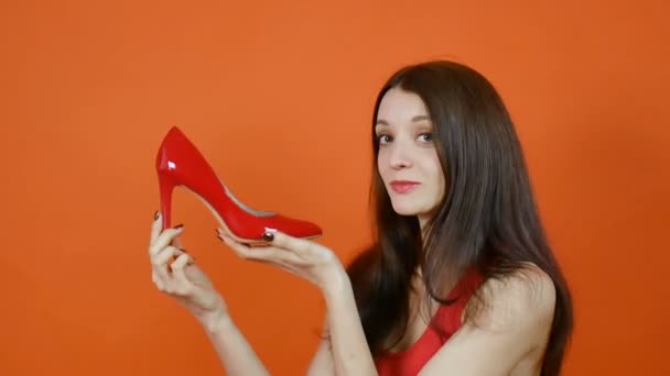 Egy szép fiatal barna hajú lány vizsgálja egy pár piros cipő. Érzelmek. Művészi portré stúdió narancssárga háttérrel - Felvétel, videó