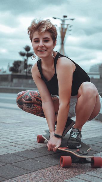 Beautiful young girl with tattoos sits on longboard - 写真・画像