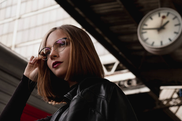 Attractive young woman millenial in black clothes and a hat and glasses at the railway station next to the train. Serious look, waiting for the train. - Foto, Bild